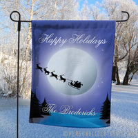 Santa's sleight flying in front of the moon above a snowy tree lined scene with your personalized greeting on a welcome yard flag
